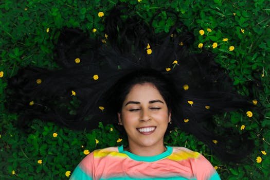 A smiling woman with closed eyes lies on green grass with yellow flowers, epitomizing serenity and happiness. cabelo crescer