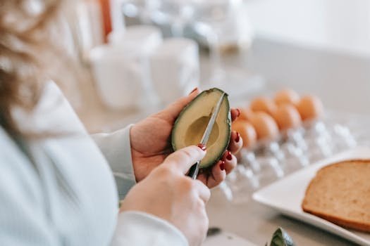 Crop unrecognizable female with knife cutting fresh avocado while making toasts at table with eggs in light kitchen during morning pele