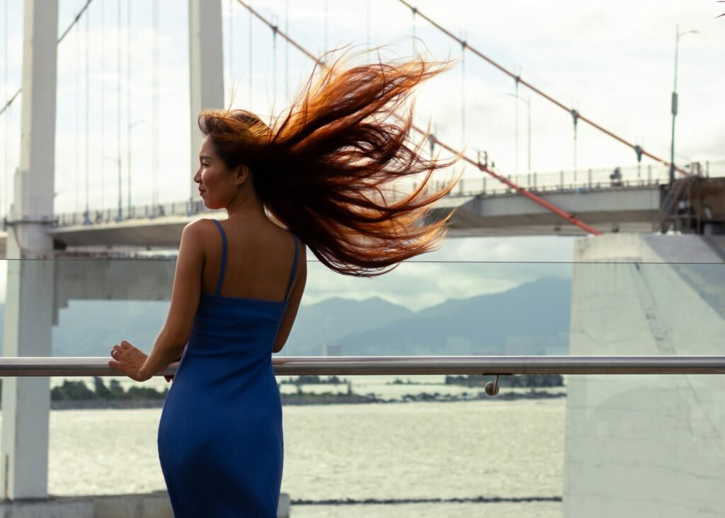 Young woman in a blue dress stands on a bridge with wind blowing her hair, overlooking water.