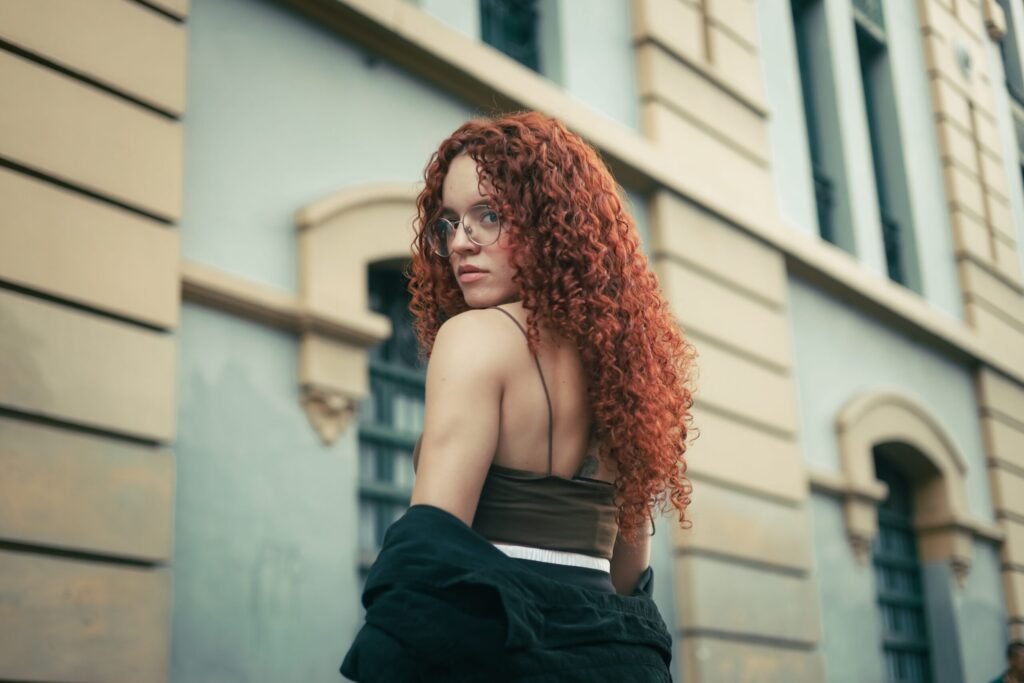 Stylish portrait of a young woman with red curls, posing outdoors in a city context.