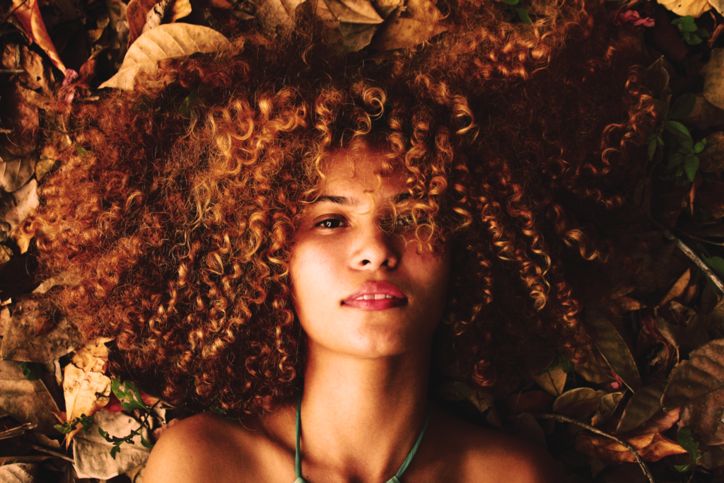 Beautiful woman with curly hair lying on dried autumn leaves, showcasing natural beauty and fall fashion.